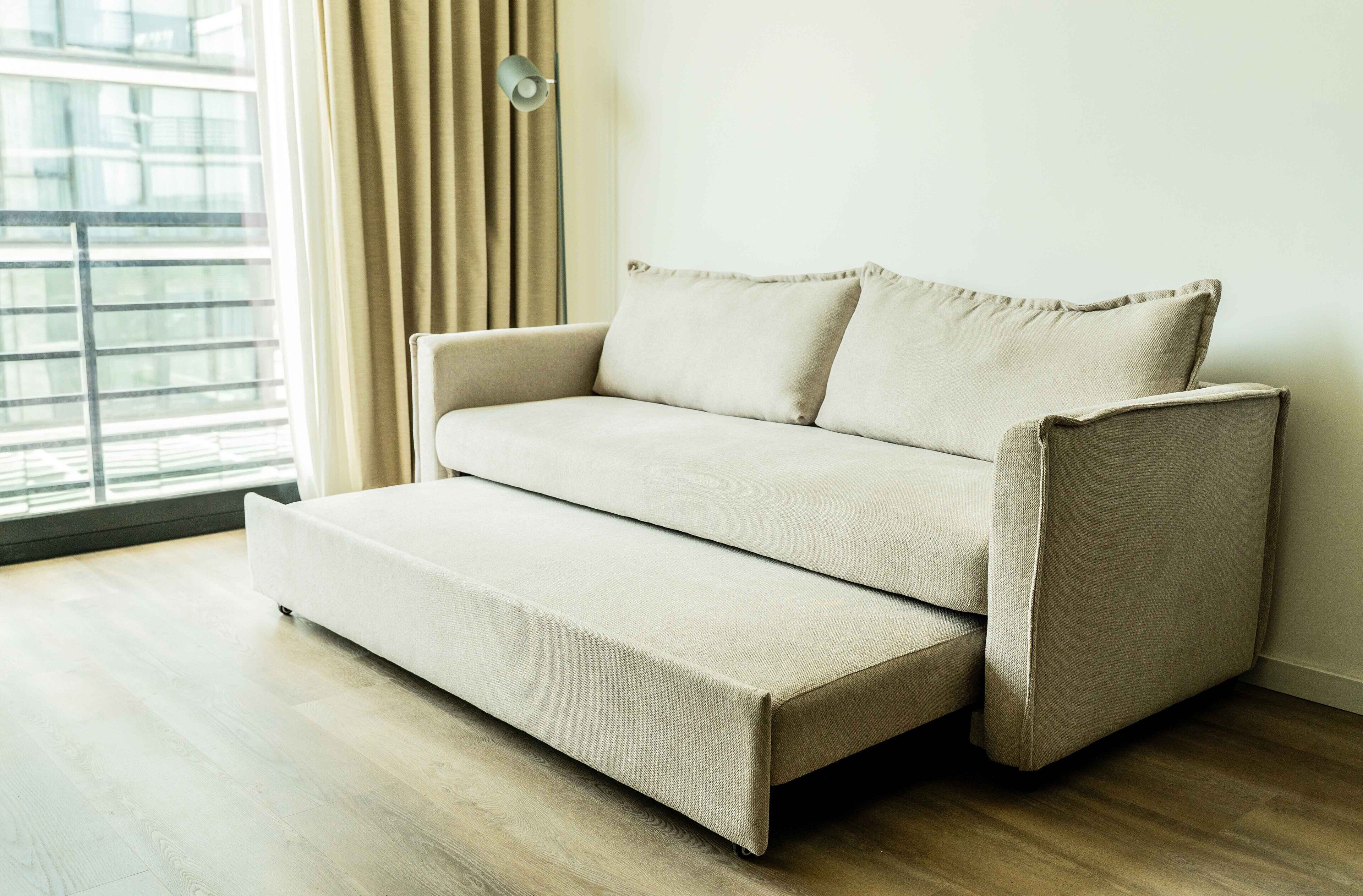 Sofá-cama num quarto com soalho de madeira e janela com cortinas.