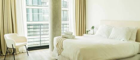 Habitación de hotel con una cama grande, una silla, una ventana con cortinas y vistas a edificios.
