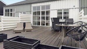 A deck with outdoor furniture, including chairs and a table, and a white building with large windows in the background. - Beautiful home in Ebeltoft with sauna (Ebeltoft)