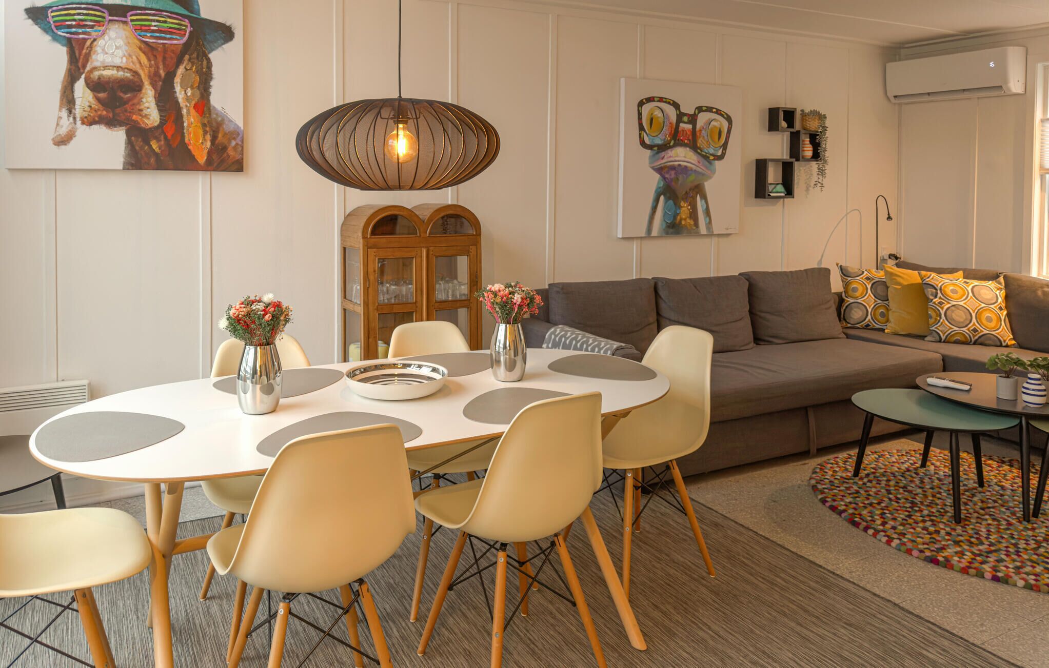 A modern living room with a round dining table, beige chairs, a sofa, and a coffee table. There are decorative vases on the table and a colorful rug on the floor.