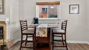 Un coin repas comprenant une table en bois, deux chaises, une machine à café et une fenêtre donnant sur un bâtiment en briques.