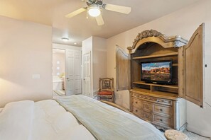 Un dormitorio con una cama, un televisor sobre un mueble de madera, un ventilador de techo y una puerta que da a otra habitación.