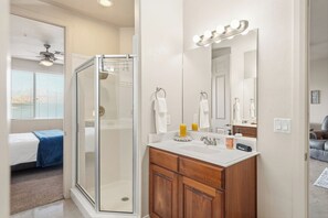 Un baño con cabina de ducha de vidrio, tocador con espejo y cama visible a través de una puerta abierta.