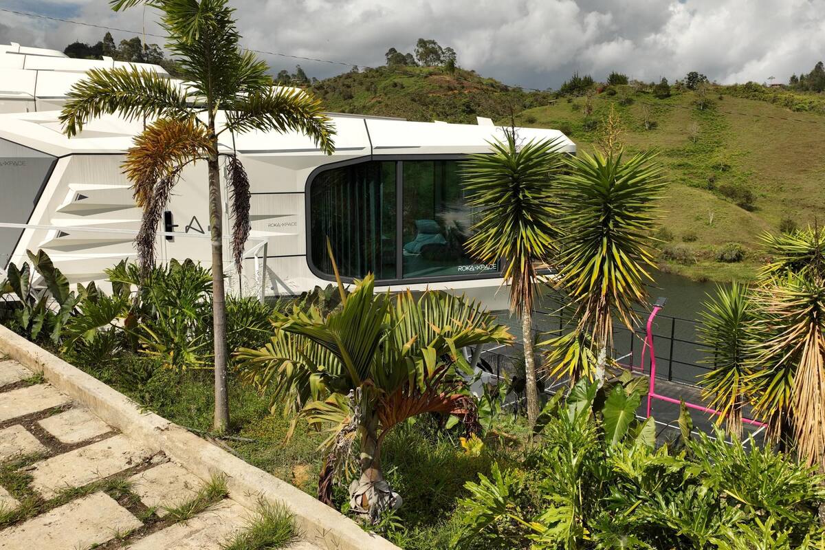 A modern RV with a curved roof, surrounded by lush greenery and a hilly landscape.
