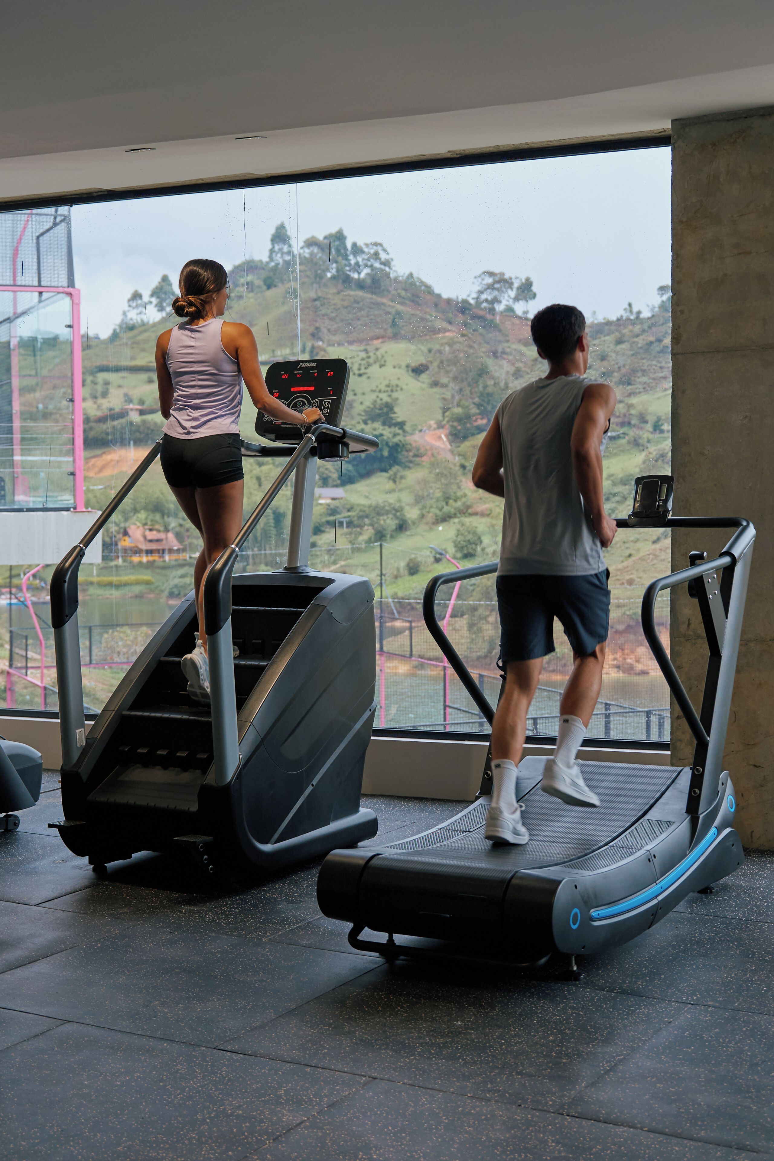 Dos personas usando cintas de correr en un gimnasio con grandes ventanales que dan a un paisaje montañoso.