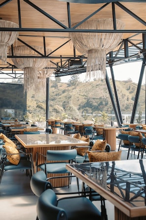 An outdoor dining area with tables and chairs, a wooden ceiling structure, and a view of a hilly landscape. - Roka Hotel (El Peñol)