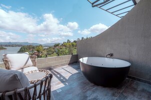 Un baño exterior moderno con una bañera independiente, sillas de mimbre y vistas a un lago y montañas.