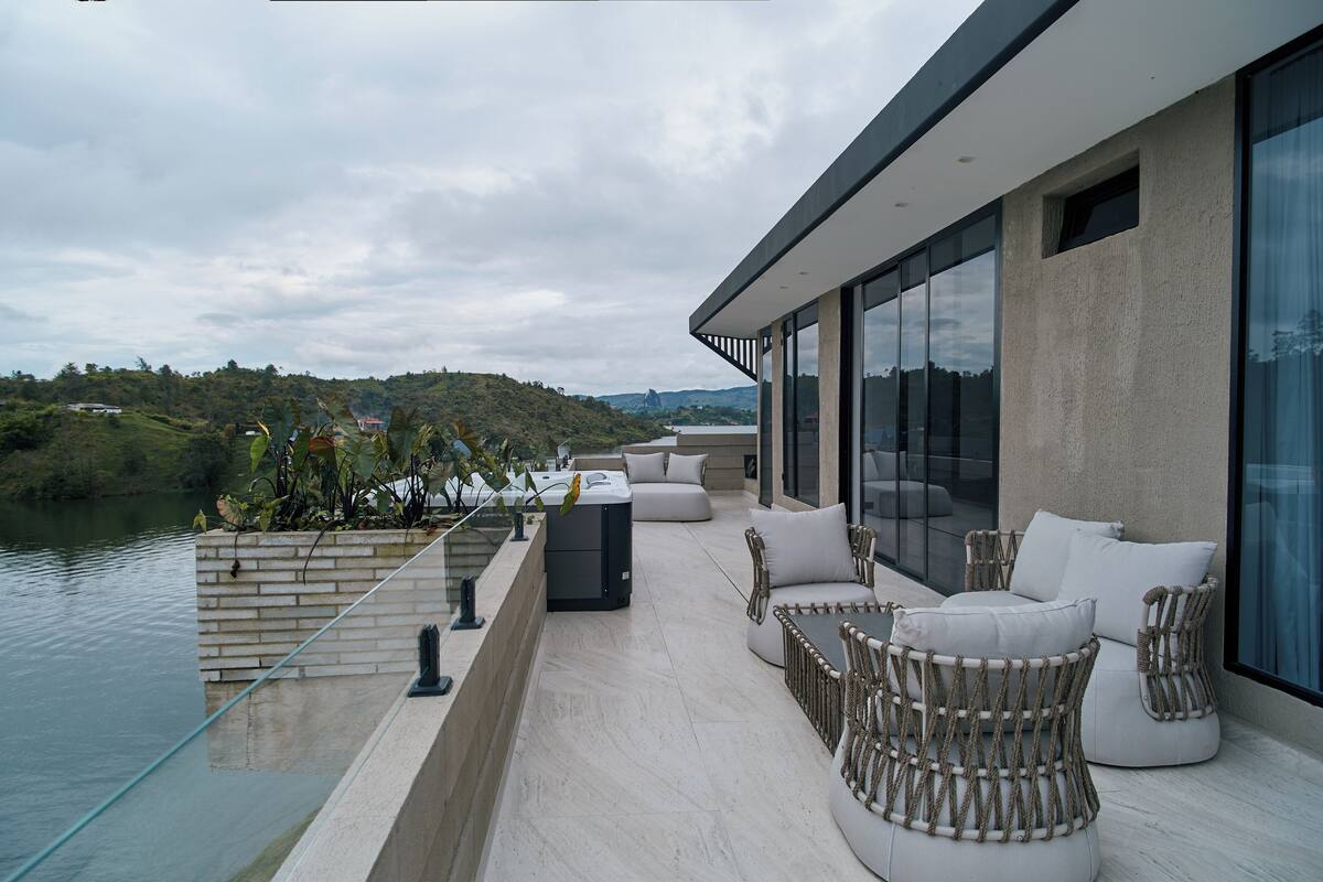A modern outdoor patio with seating, a glass balustrade, and a view of a river and hills.