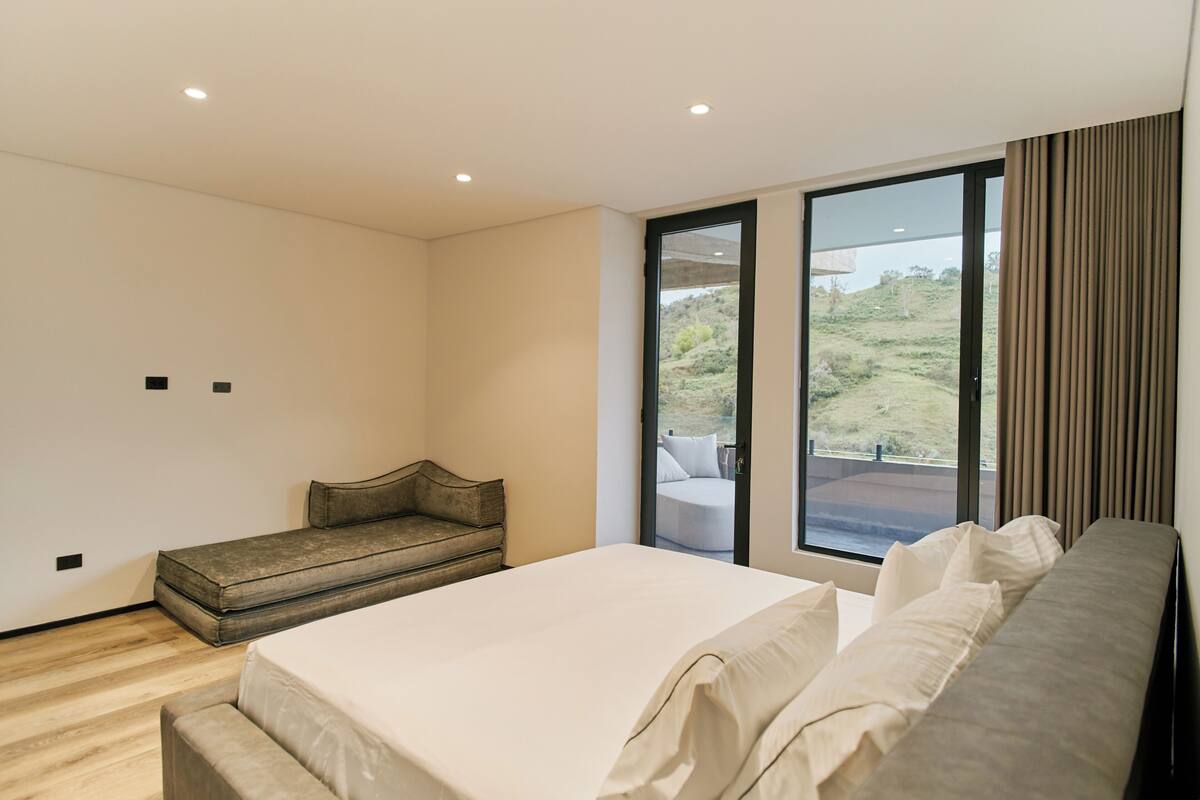 A bedroom with a bed, a bench, and a large window offering a view of the outdoors.
