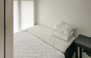 Una cama bien tendida con una colcha blanca acolchada y dos almohadas, una mesita de noche pequeña y una pared lisa.
