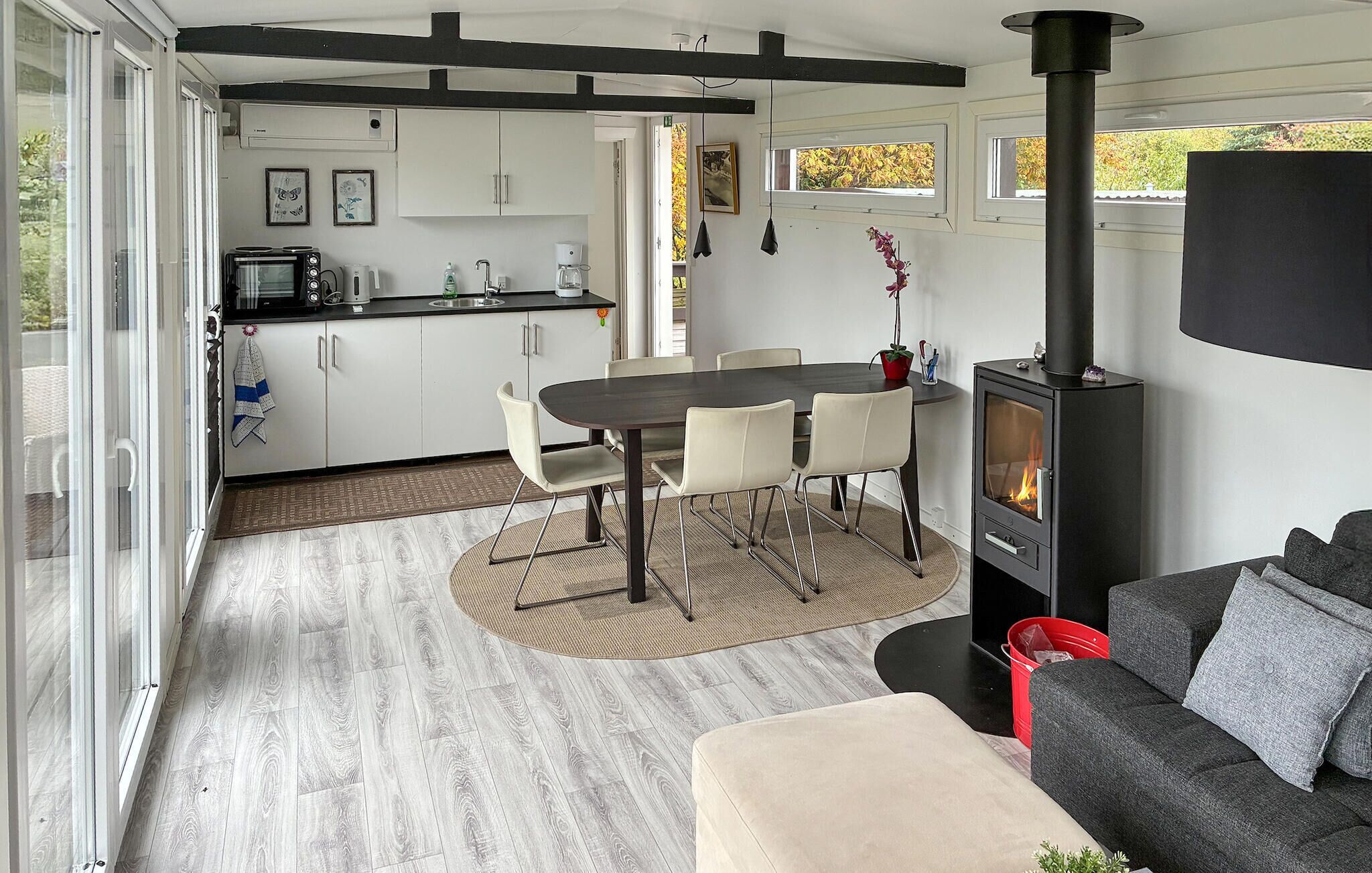 A modern kitchen with white cabinets, a dining area with a round table and chairs, a built-in wood stove, and a grey sofa in a bright living space with large windows.