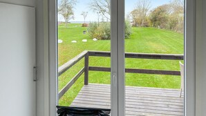 A view from a room with a sliding glass door to a deck. The deck has a wooden railing and a grassy lawn with trees and a distant view of the sea. - 2 bedroom amazing home in Samsø (Samsø)