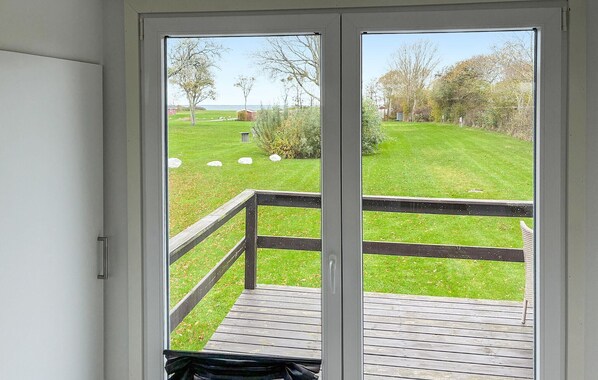 Vista desde una habitación con puerta corrediza de vidrio a una terraza. La terraza tiene baranda de madera y un césped con árboles, y una vista distante del mar.