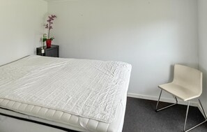 Una cama bien hecha con colcha blanca, una mesita de noche con un jarrón rojo y una orquídea, y una silla sencilla en una habitación minimalista.