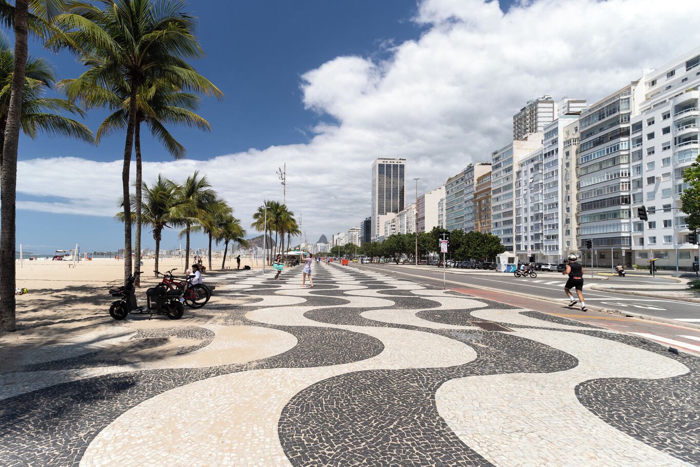 A beachside promenade with palm trees, a patterned pavement, and modern buildings in the background.