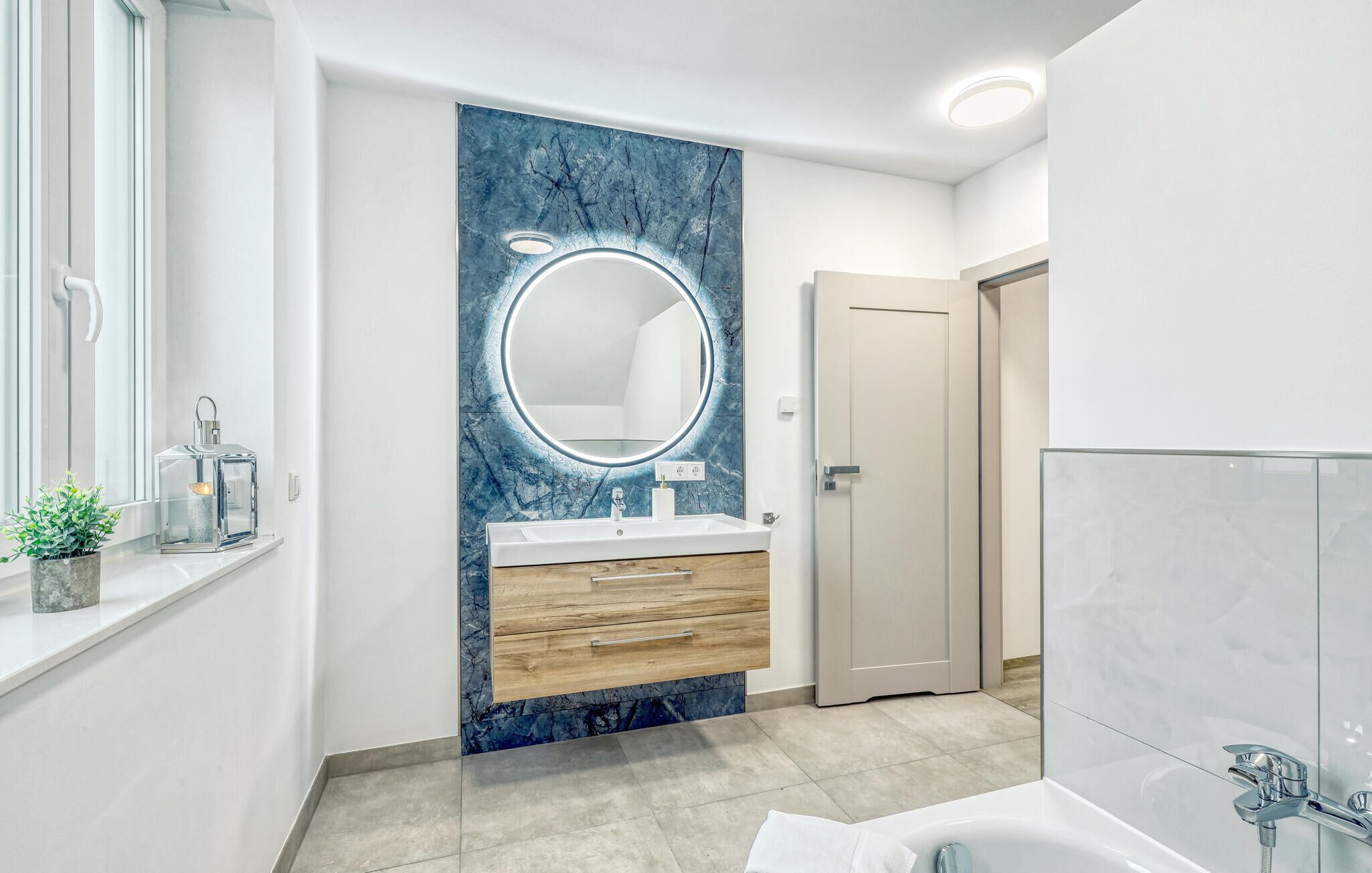 Un baño moderno con tocador de mármol, espejo redondo, lavamanos blanco y bañera.