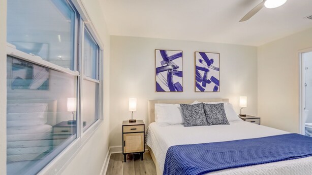 Una habitación de hotel con una cama grande, dos mesitas de noche con lámparas, un ventilador de techo y dos cuadros abstractos en la pared.