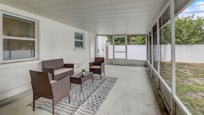 Un patio cubierto con mobiliario de mimbre, un tapete estampado y grandes ventanales que ofrecen vistas al exterior.