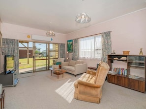 A spacious living room with a sofa, armchair, coffee table, and a bookshelf. - Hosts on the Coast Cosy on Cooks (Cooks Beach)