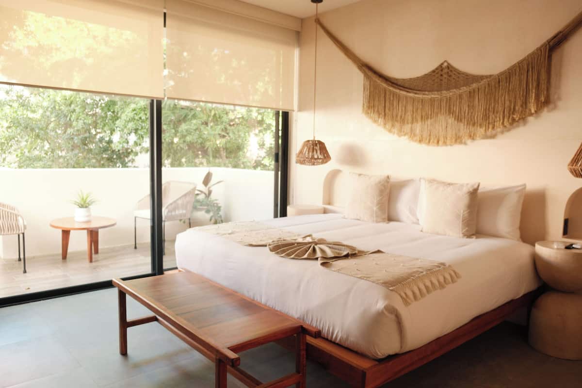 Een slaapkamer met een groot bed, een bankje, een klein tafeltje en een macramé wanddecoratie.