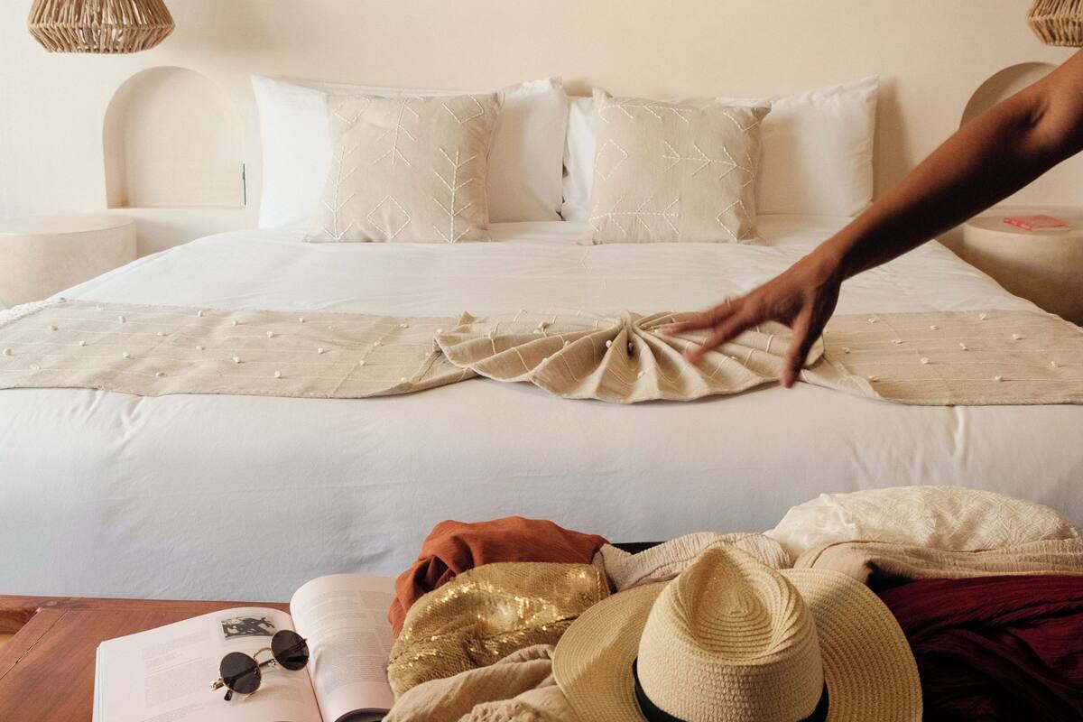Een bed met een macramé wanddecoratie, een nachtkastje met een boek en een bril, en een strohoed op de vloer.