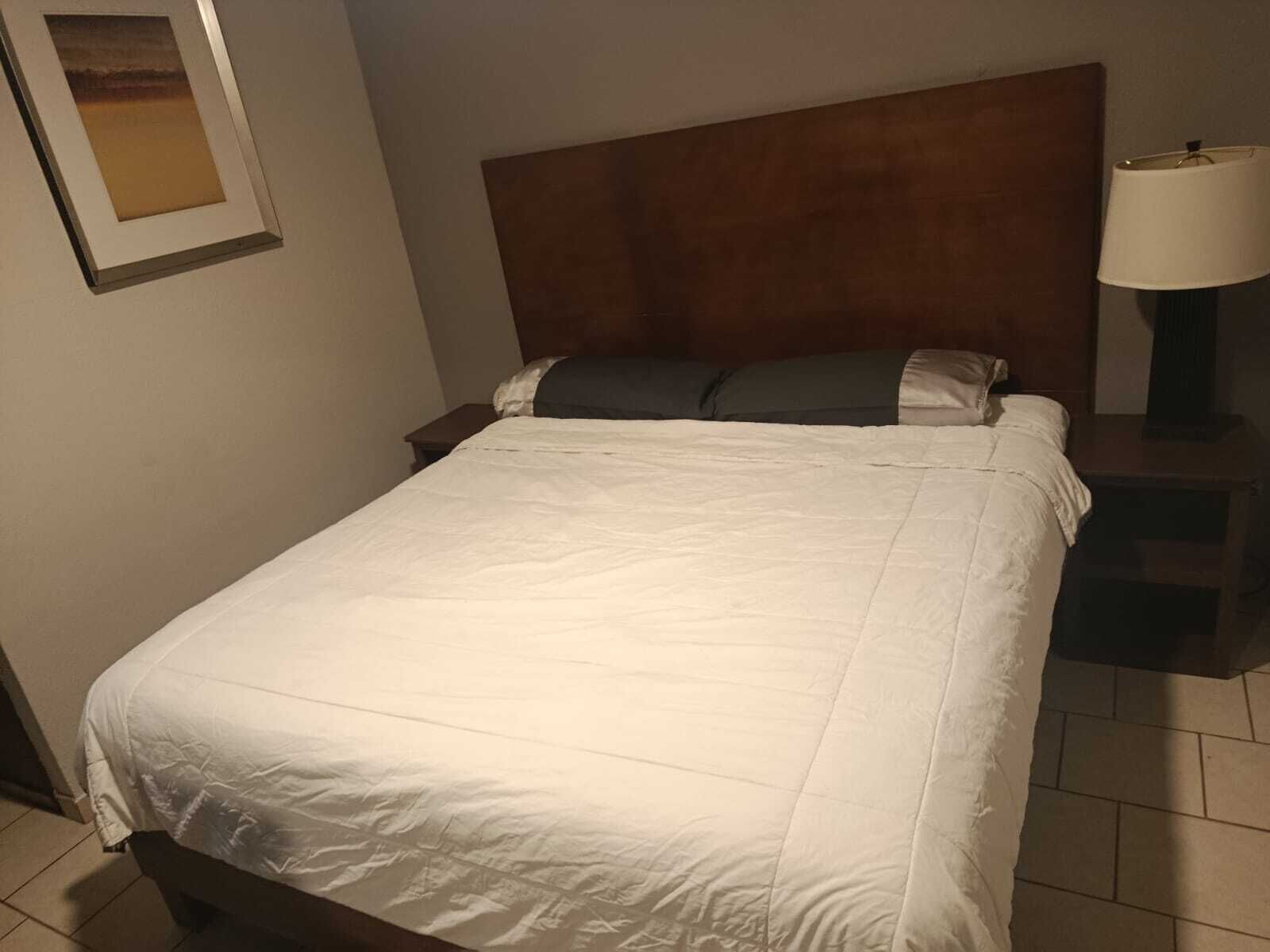A neatly made bed with a white duvet, a wooden headboard, and a nightstand with a lamp.