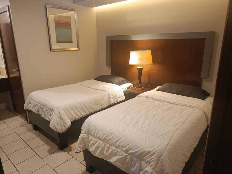 Quarto de hotel com duas camas, uma mesa de cabeceira com abajur, um quadro na parede e uma cabeceira de madeira.