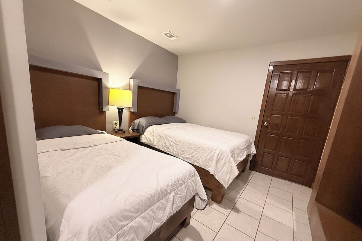 A hotel room with two single beds, a nightstand with a lamp, a wooden door, and tiled flooring.