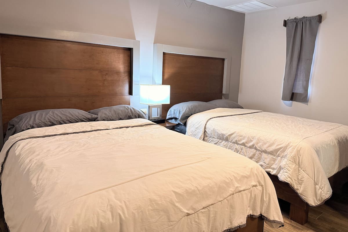 A hotel room with two beds, a bedside table with a lamp, and a window with curtains.