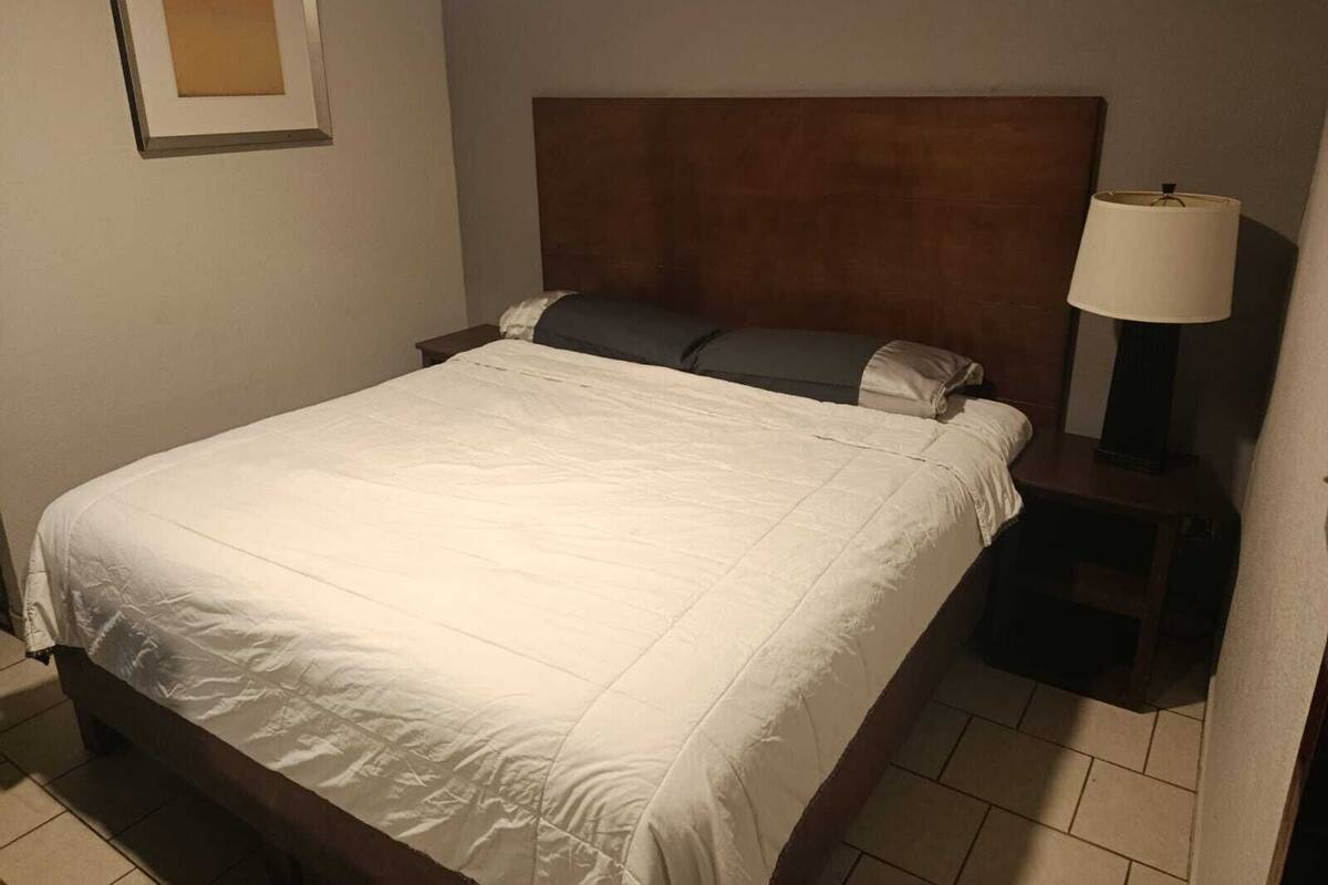 A neatly made bed with a white comforter and pillows, a wooden headboard, a bedside table with a lamp, and a framed picture on the wall.