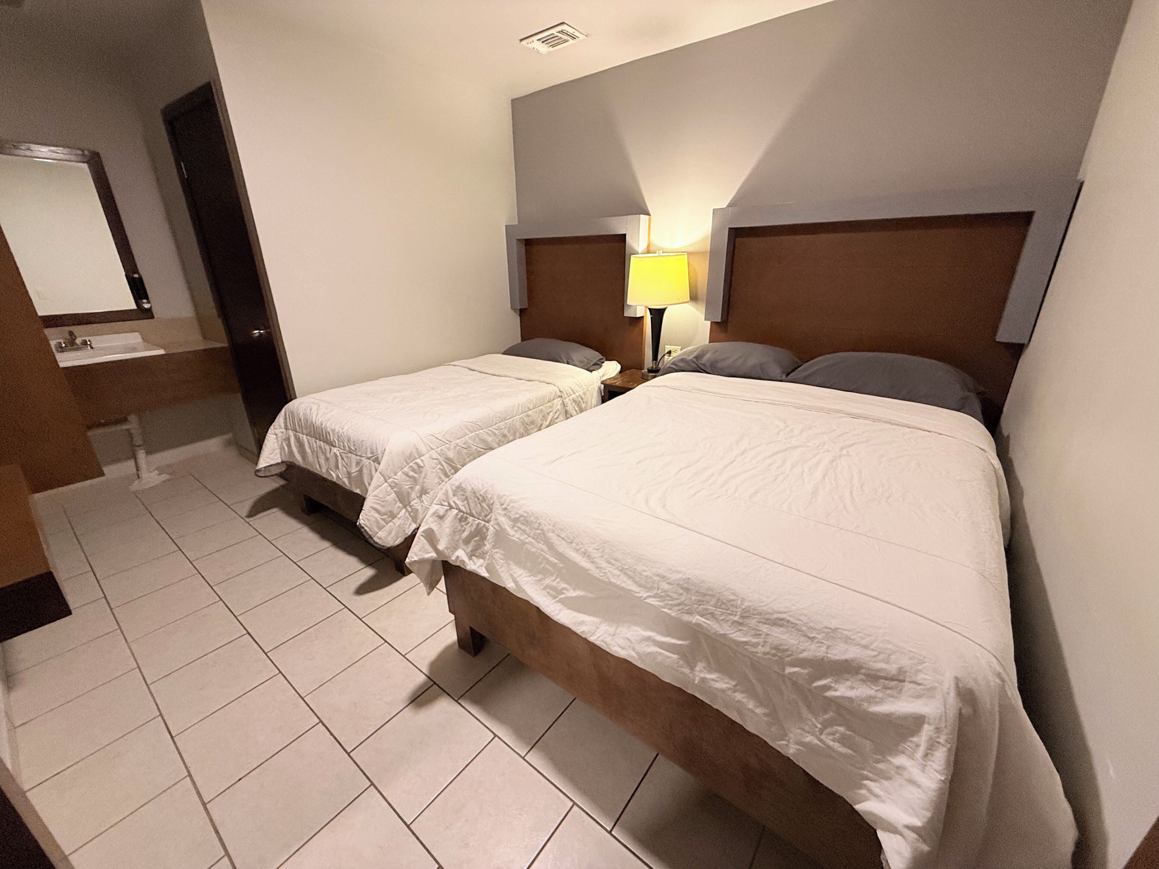 A hotel room with two single beds, a bedside table with a lamp, a mirror, and a bathroom with a sink and toilet.
