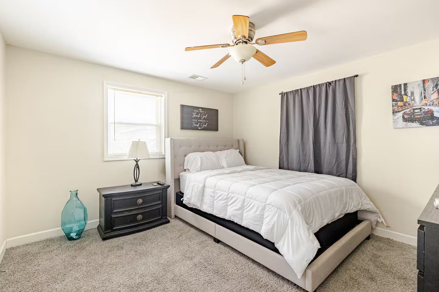 A neutral bedroom with an upholstered bed, ceiling fan, and carpeted flooring.