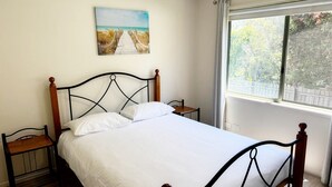 A neatly made bed with white linens, a wooden headboard, and two nightstands with a small table on each. A painting of a beach scene hangs above the bed. - Tanjil Cottage | Relaxing Coastal Stay (Lakes Entrance)