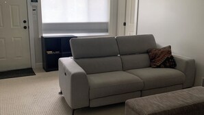 A living room with a beige sofa, a matching ottoman, a window with blinds, a door, and a ceiling fan.