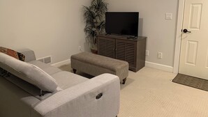 A living room with a grey sofa, a grey ottoman, a television on a cabinet, and a potted plant.
