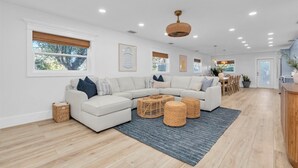 A modern living room with a sectional sofa, wicker ottomans, and a dining area with wooden chairs. - “Aura” 5min to beach- sleeps 20- pool with waterfall and fire features (Bradenton)