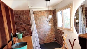 A compact room with a brick wall, a wooden floor, a window with curtains, a hanging light fixture, and a small table with a blue container. - Secret travel lodge, Au Calme Absolu (Lindry)