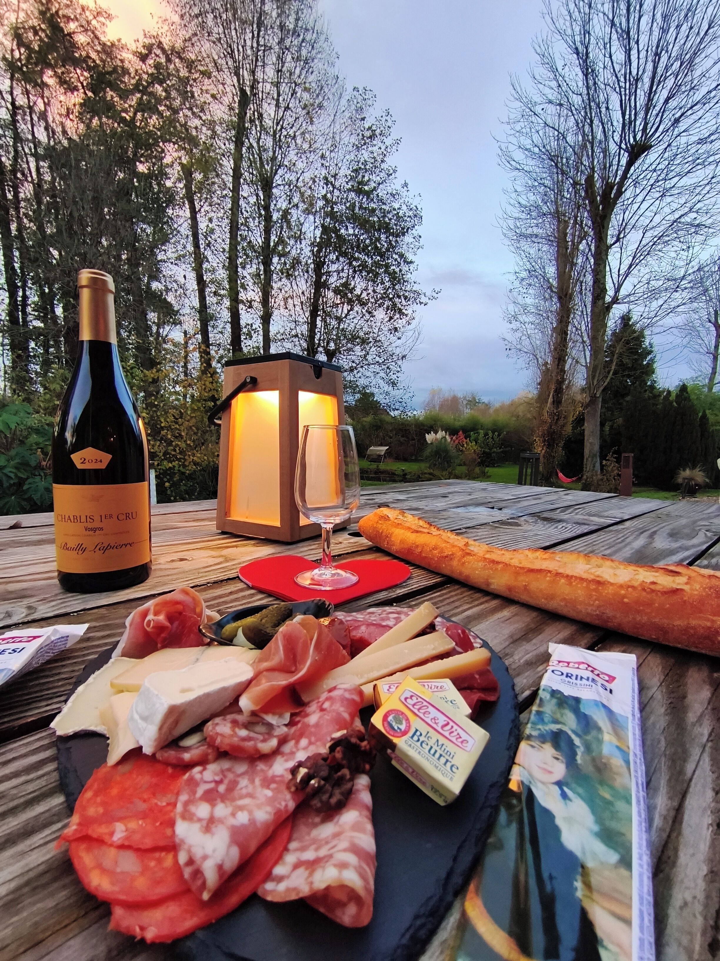 Un pique-nique installé sur une table en bois à l’extérieur, avec une bouteille de vin, un verre, une baguette, divers fromages et de la charcuterie.