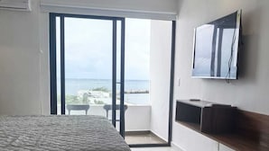 Ein Zimmer mit einem Bett, einem Tisch für zwei Personen, einem Sessel, einem an der Wand montierten Fernseher und einem Balkon mit Meerblick.