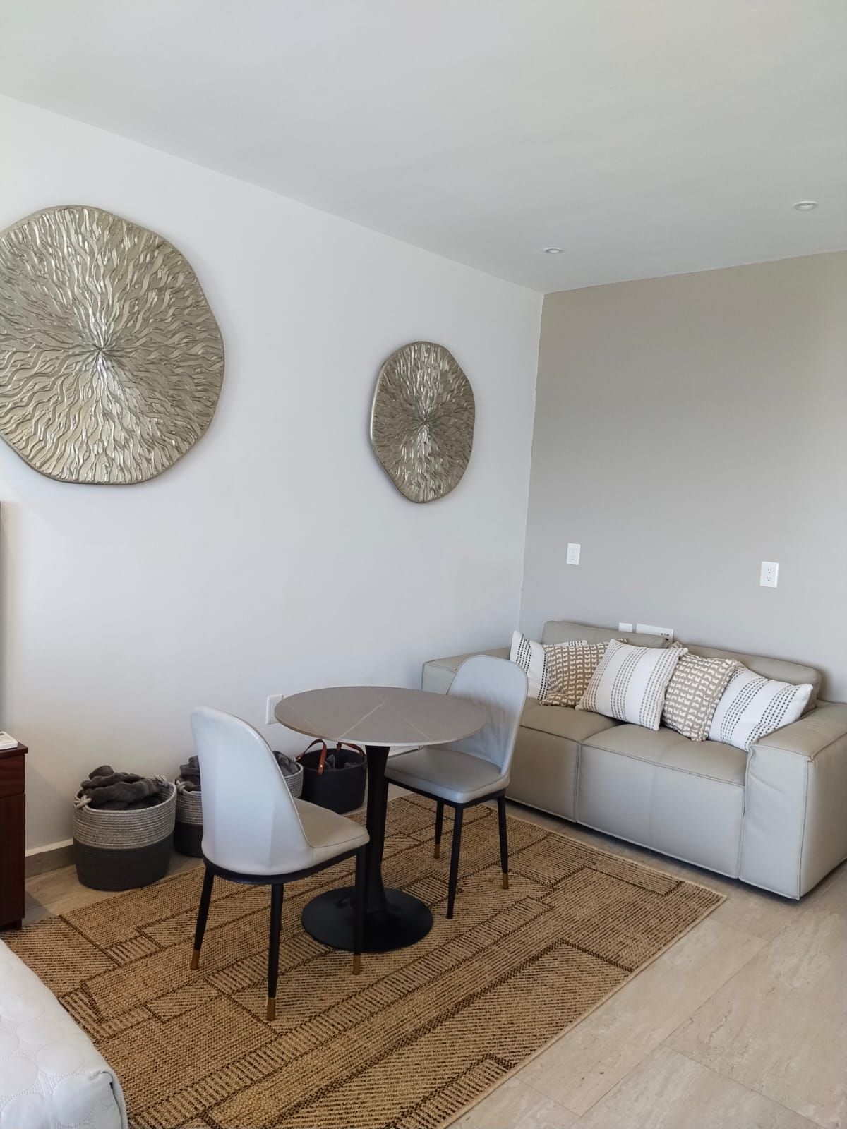 A living room with a round table, two chairs, a sofa, and two wall art pieces.