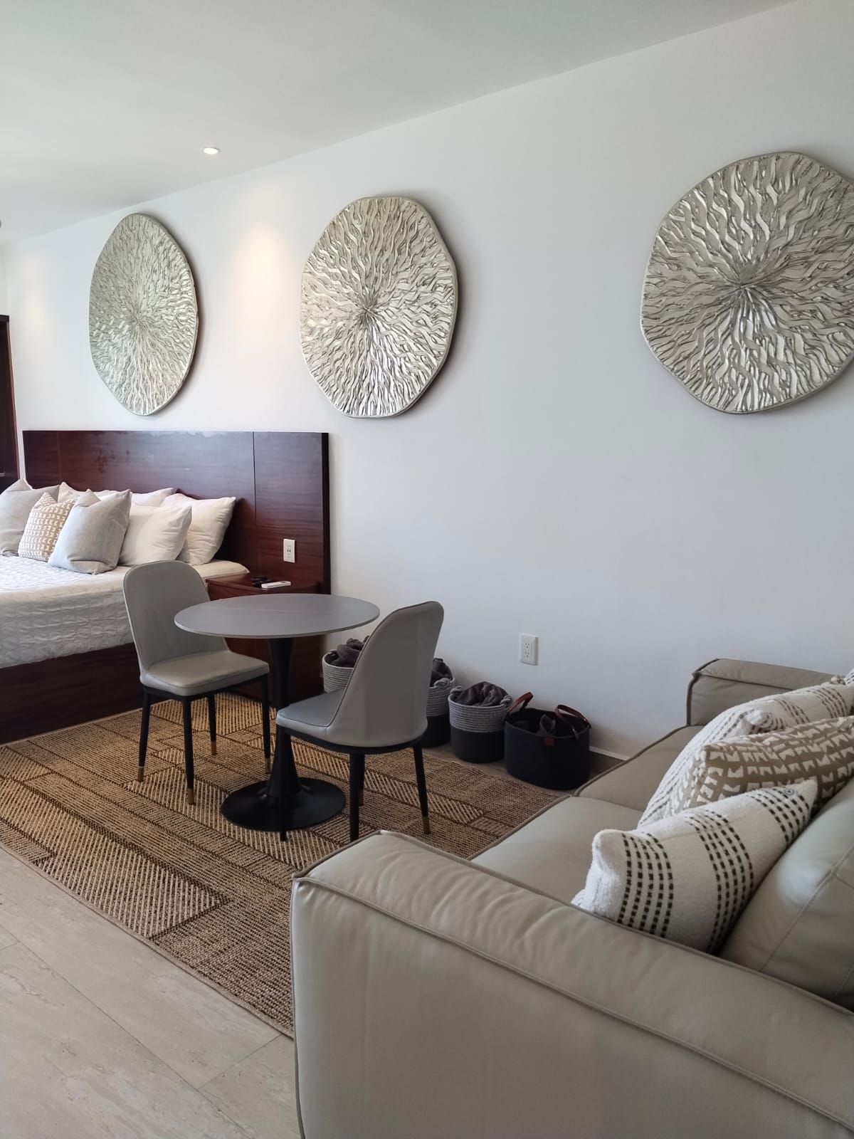 Habitación de hotel moderna con una cama, una mesa de comedor redonda, dos sillas, un sofá y tres adornos en la pared.