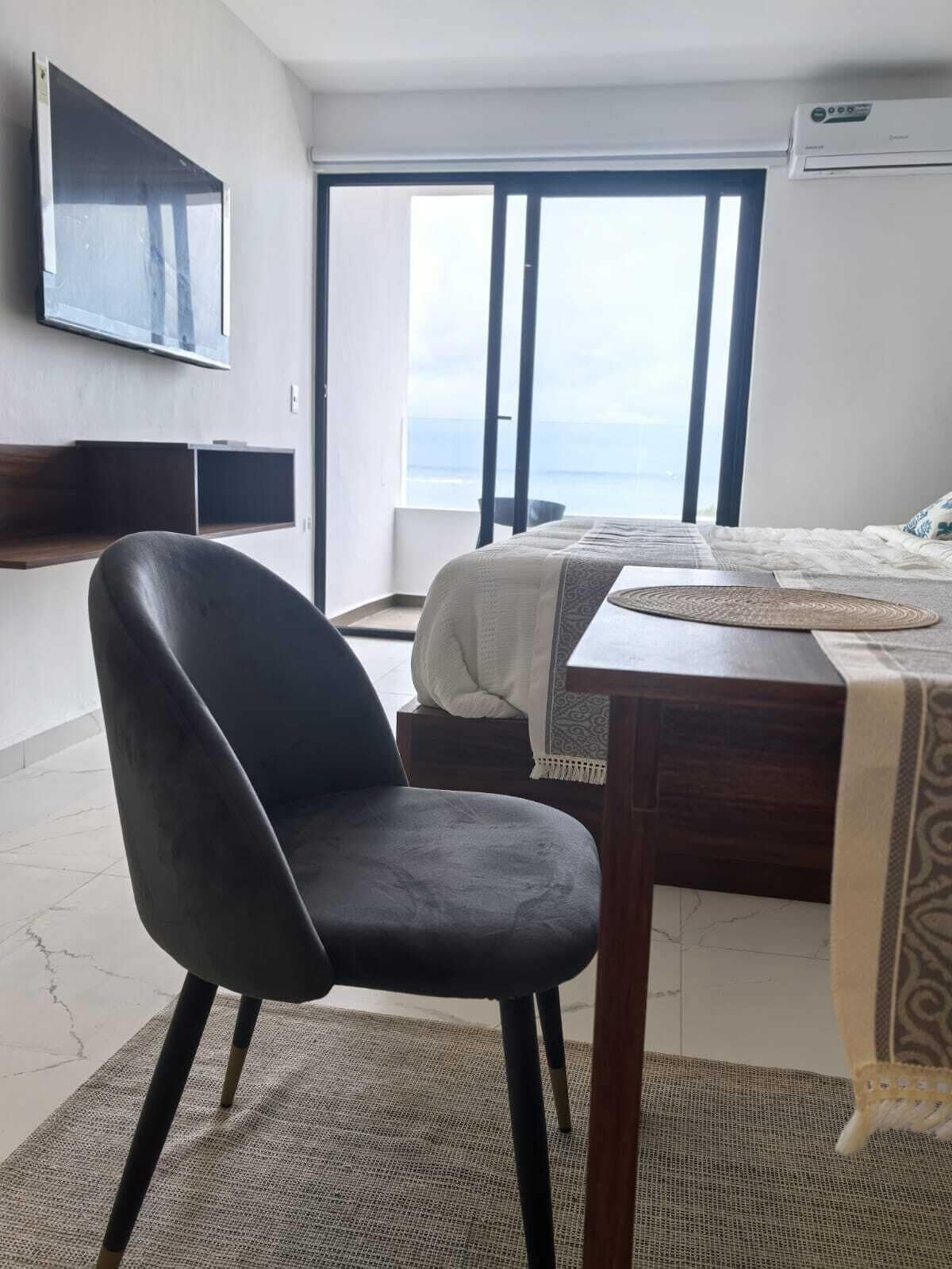 A modern hotel room with a large window, a flat-screen TV mounted on the wall, a wooden desk, and a comfortable chair.