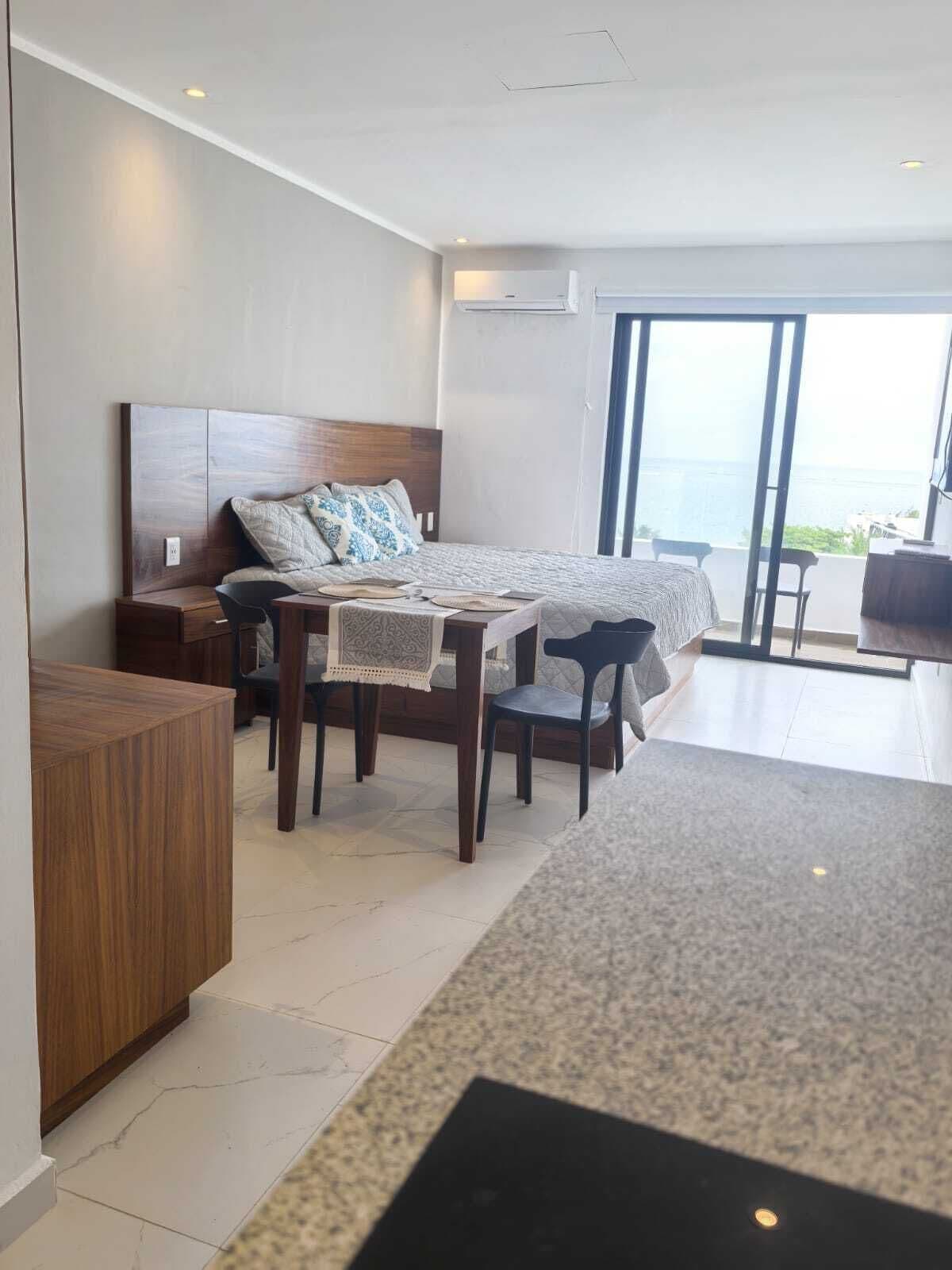 A modern bedroom with a large bed, a desk, and a chair. There is a view of the ocean through the window.