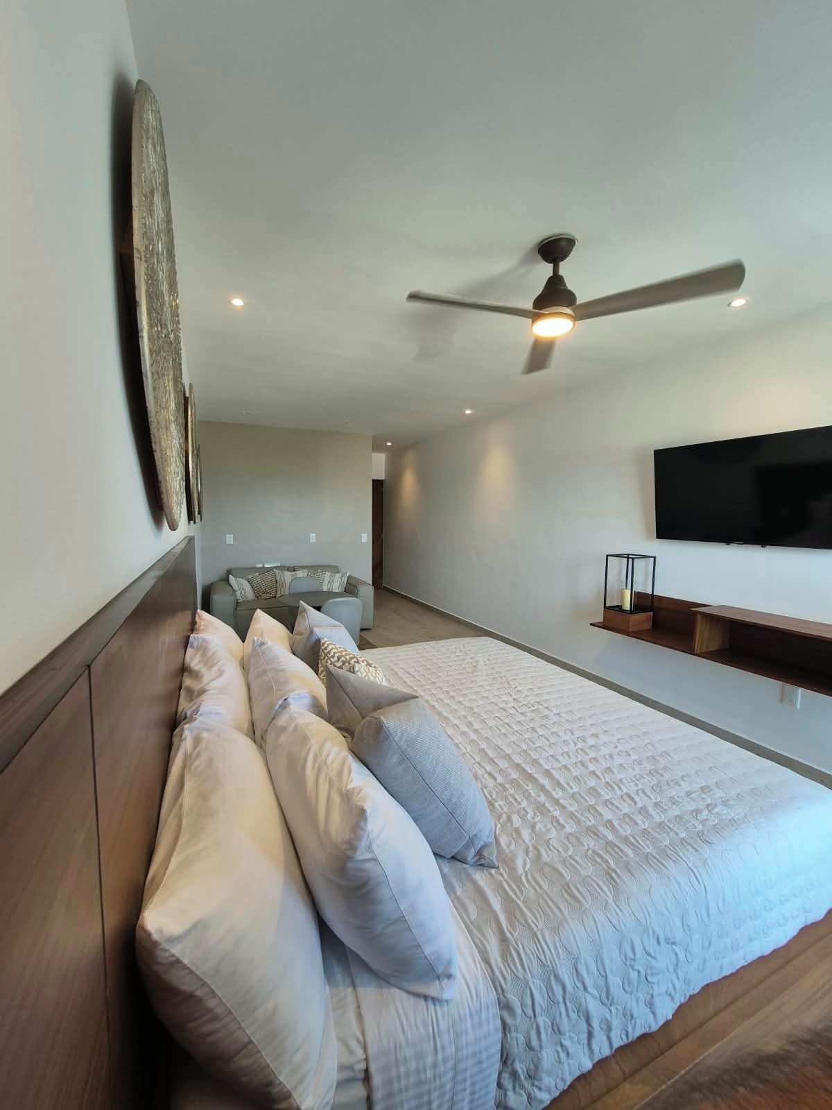 A modern bedroom with a large bed, a ceiling fan, a flat-screen TV, and a seating area with a sofa and a coffee table.