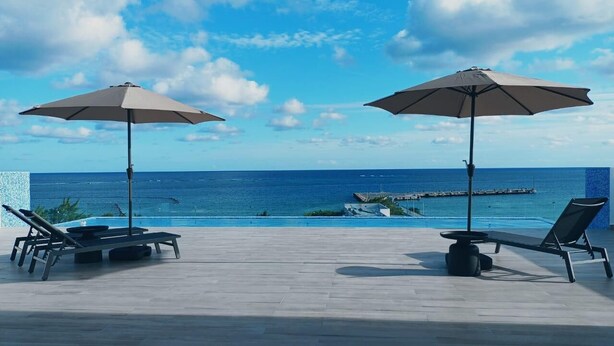 Eine Terrasse mit zwei Liegestühlen und Sonnenschirmen mit Blick auf das klare, blaue Meer und einen Pier in der Ferne.