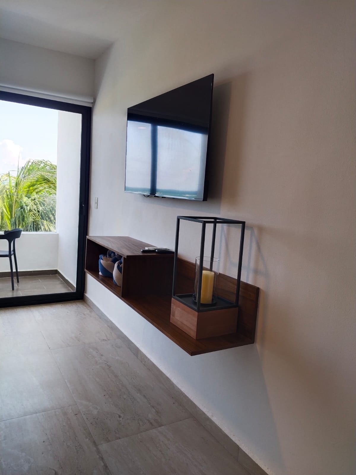 Una sala de estar moderna con un televisor de pantalla plana montado en la pared, un estante de madera con una vela, y vistas al exterior a través de una puerta corrediza.