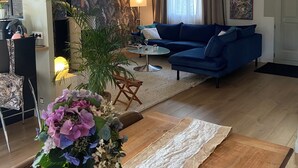 A modern living room with a blue sofa, a dining table with a lace table runner, and a large window with curtains. - Charming single-storey house (Évran)