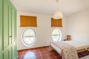Een kamer met een bed, twee ramen met zonwering, een hanglamp en een groene deur.