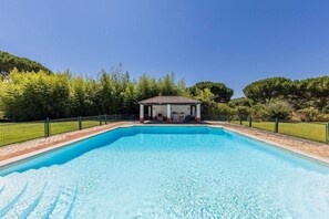 A rectangular swimming pool with a covered pavilion in the center, surrounded by a fence and trees.