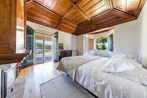 A bedroom with a large bed, a wooden ceiling, a view of the outdoors, and a door leading to a balcony.